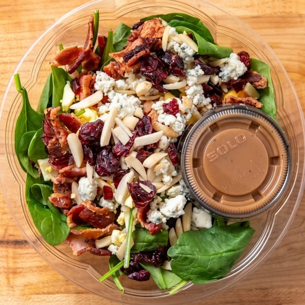 Spinach salad with bacon, blue cheese, cranberries, almonds, and dressing in a clear plastic container.